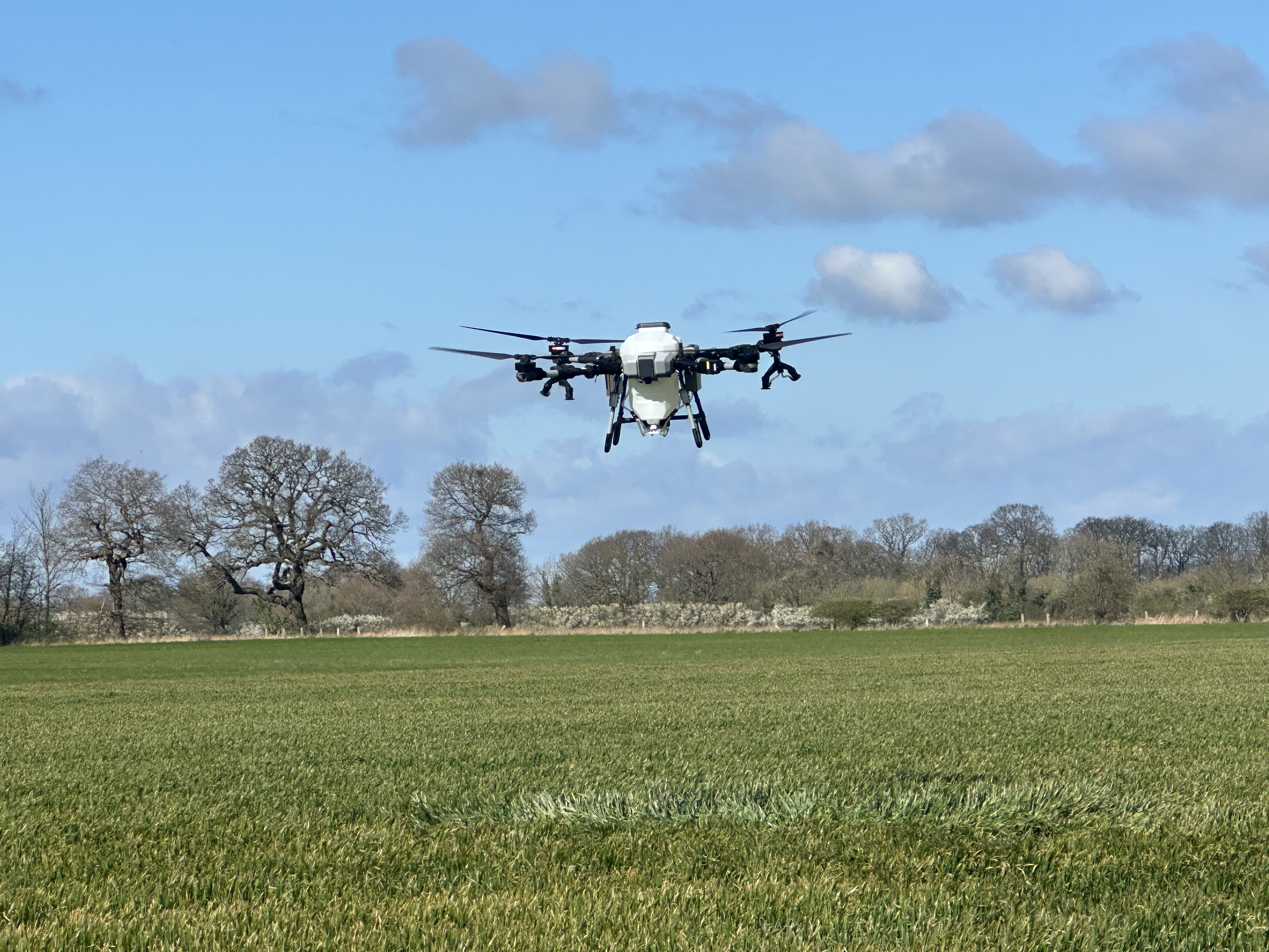 FP700 application drone hovering above a green cereal crop under blue sky
